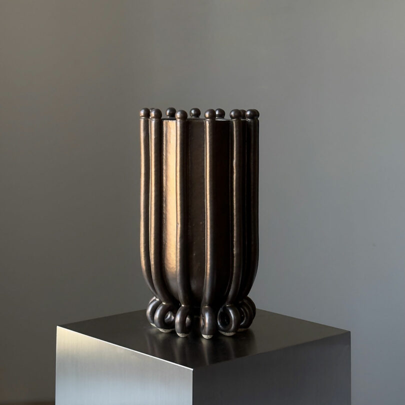 A tall, dark vase with vertical ridges and a metallic sheen sitting on a pedestal.