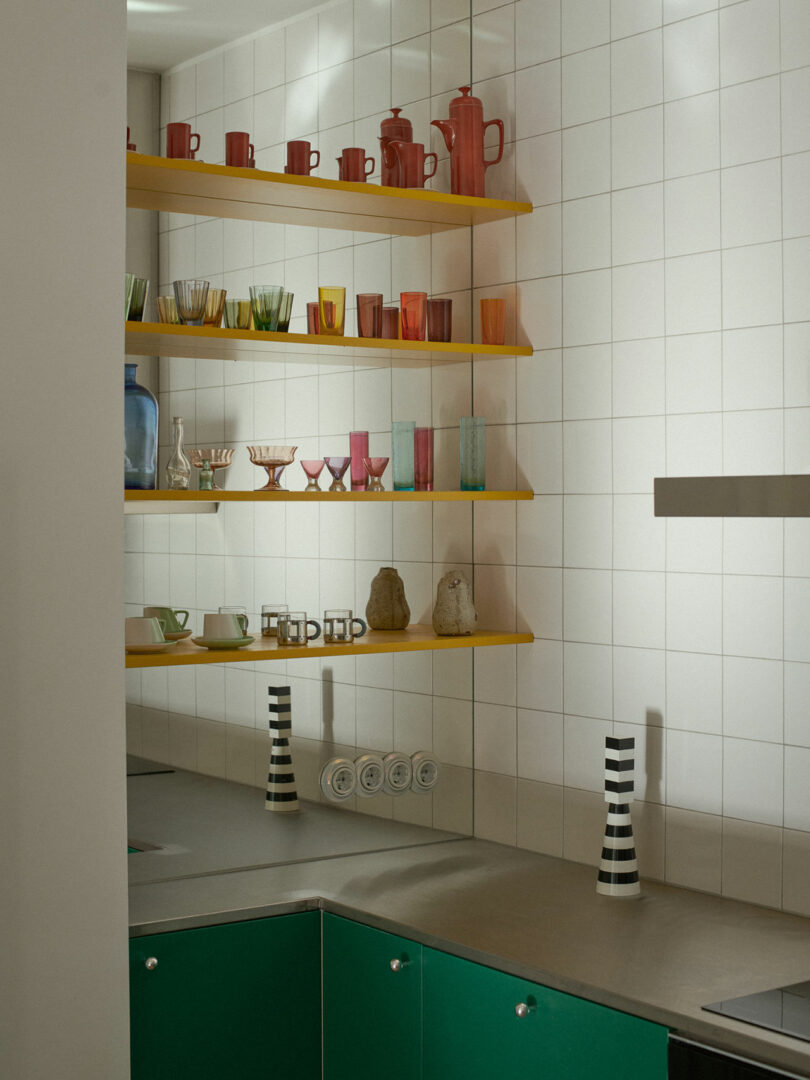 Detail of green Valchromat kitchen cabinets with yellow shelving and colorful glassware.