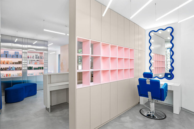 Interior view featuring an illuminated wavy mirror, pink shelving, and a signature cobalt blue chair.