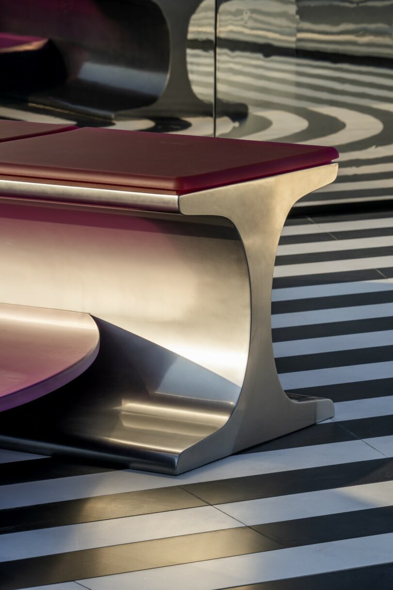 A modern stool with a red cushion and a reflective metallic base set against a striped floor.