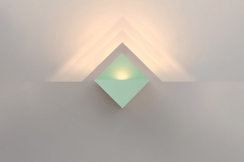 Geometric wall light casting triangular shadows and soft yellow illumination on a light-colored wall.