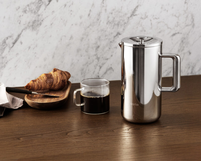 A breakfast table setup featuring the Aarke press, a cup of coffee, and a croissant on a wooden tray.