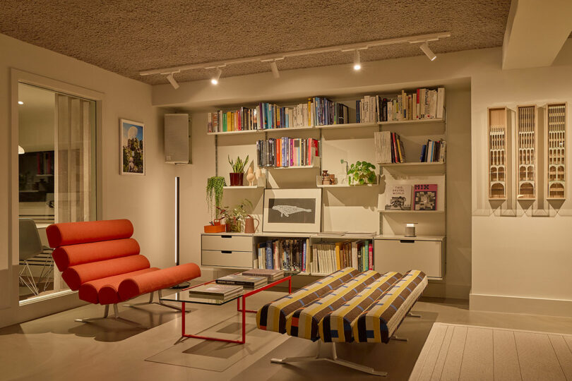 A stylish office lounge area featuring a red lounge chair, a glass coffee table, and shelves filled with books and architectural models.
