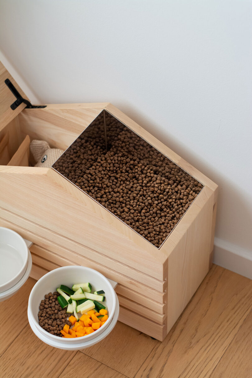A wooden container of dry food next to a white ceramic bowl containing fresh vegetables and kibble.