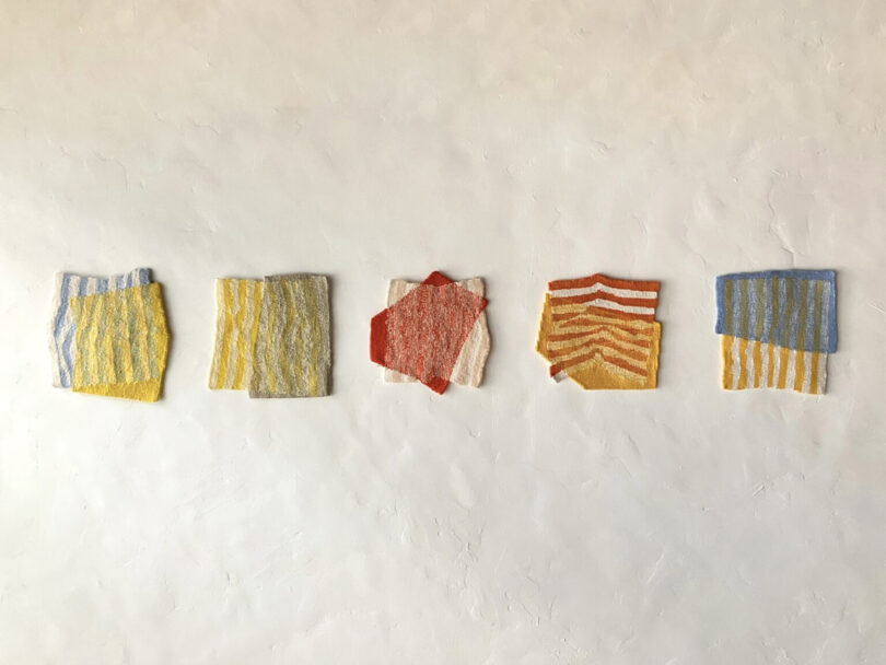 Five square fabric swatches in various colors and striped patterns arranged on a gallery wall.