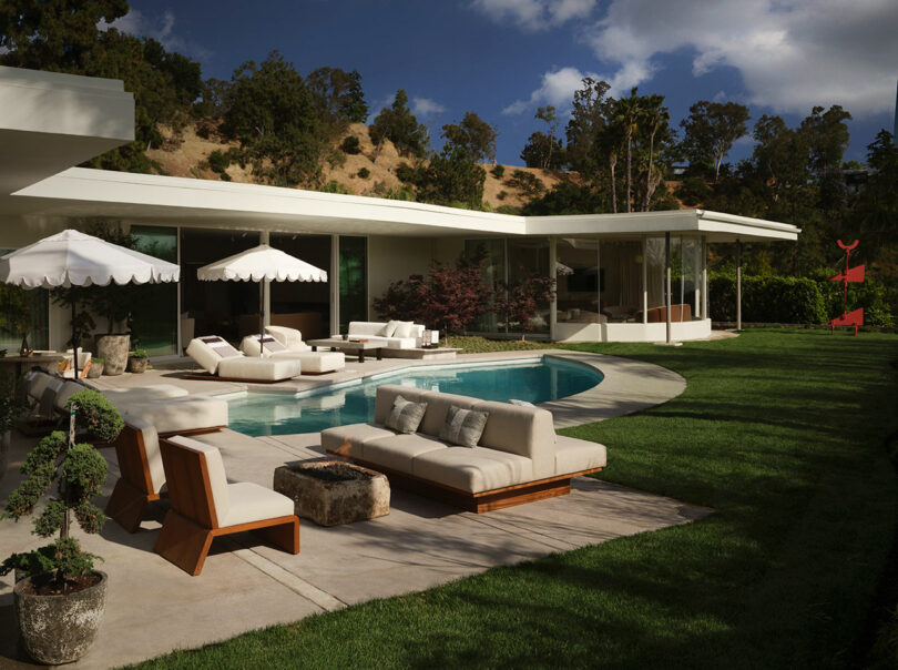A wide shot of a modern house with a swimming pool, lounge chairs, and palm trees against a mountain backdrop.