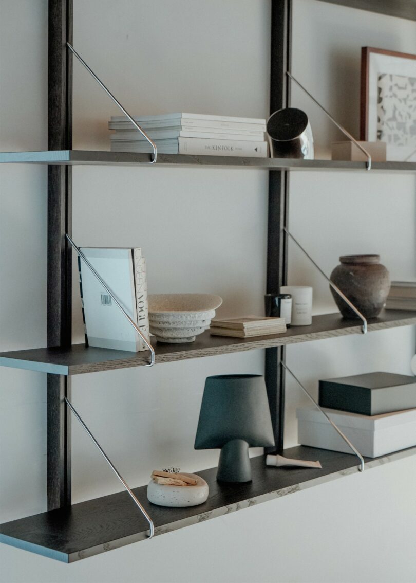 Three minimalist wooden shelves supported by thin black metal rods, arranged with books and ceramics.