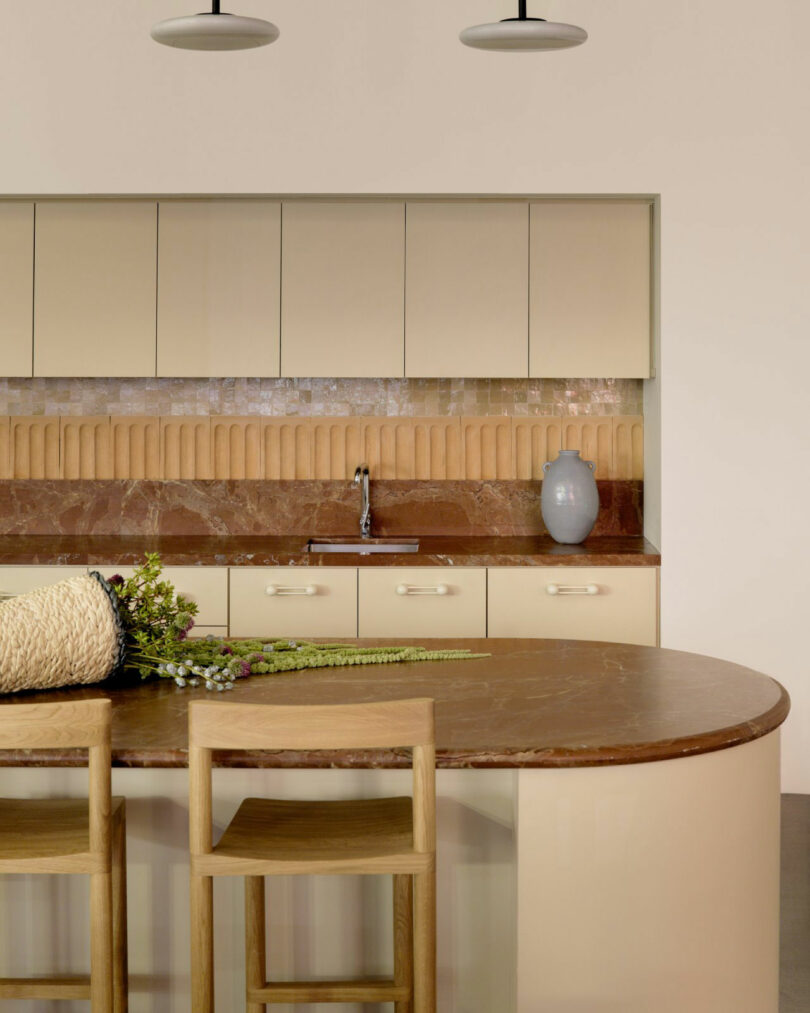 Minimalist office kitchen with beige cabinetry, brown marble island, and wooden stools under modern pendant lights.