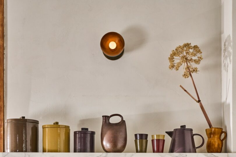 A shelf with various ceramic containers and a jug arranged beneath a wall-mounted round light.
