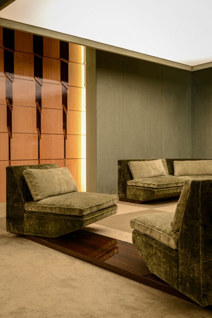 A modern lounge interior featuring olive green velvet seating and wall-integrated wooden shelving under soft architectural lighting.