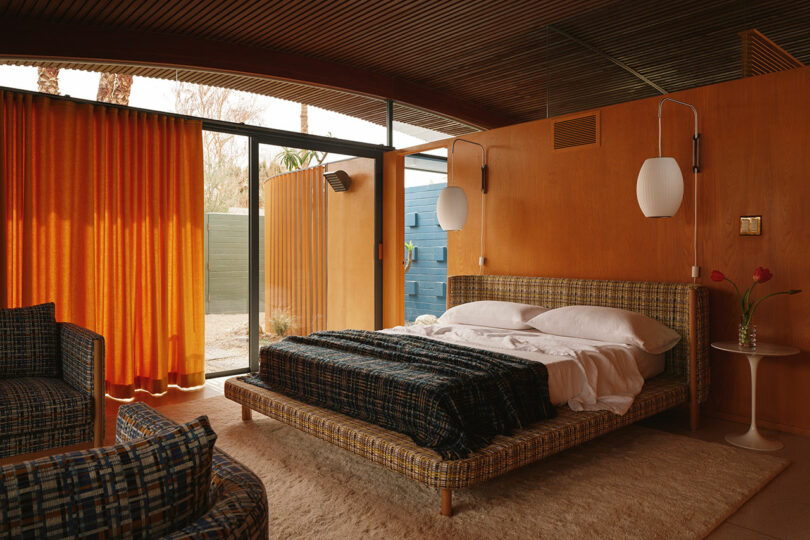 A mid-century modern bedroom with orange curtains and wood paneling leading to an outdoor space.