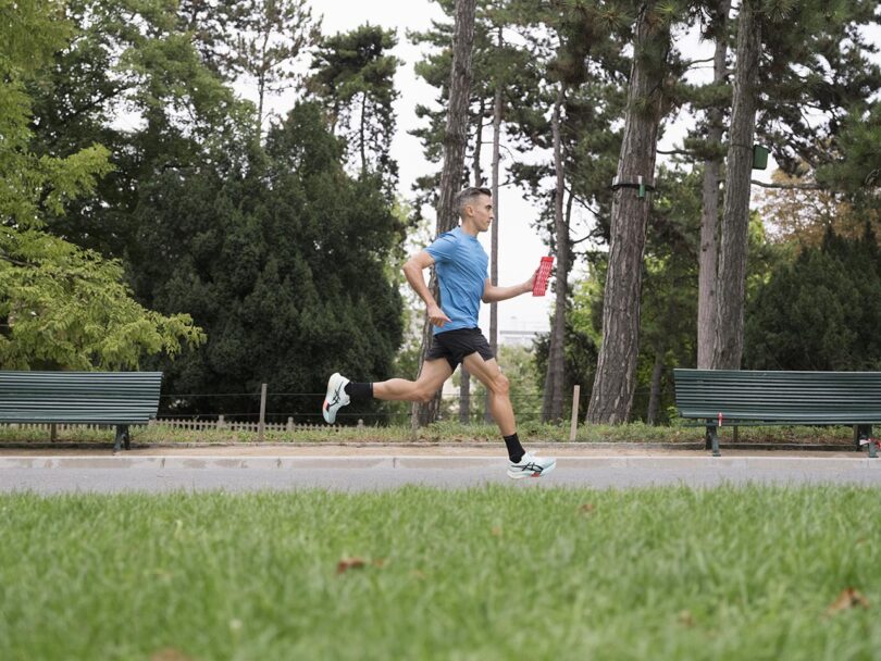 A man running through a park holding a water bottle with green park benches in the background.