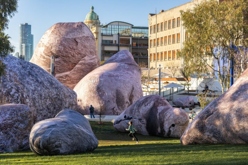 Large pink inflatable boulders scattered across a grassy park with people walking among them.