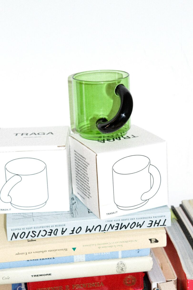 A green glass mug stacked on top of branded TRAGA boxes and coffee table books.