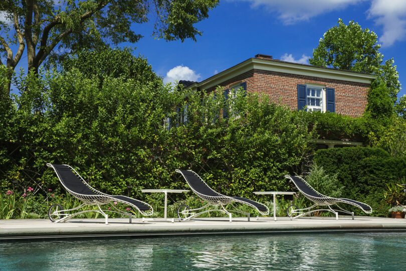 Three modern lounge chairs and side tables set against a swimming pool and a classic brick house.