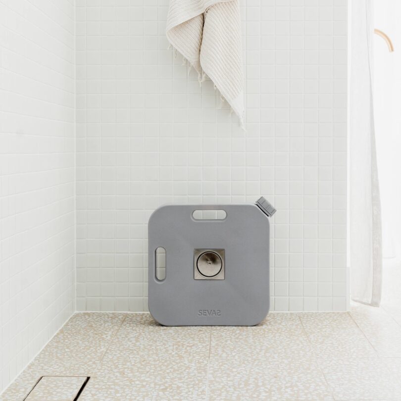 A gray square SEVAS water container with a spout placed on a tiled bathroom floor.