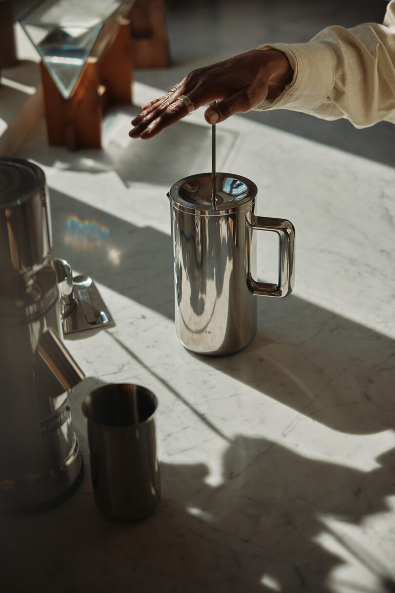 A hand pressing down the plunger of the stainless steel coffee maker on a white marble countertop.