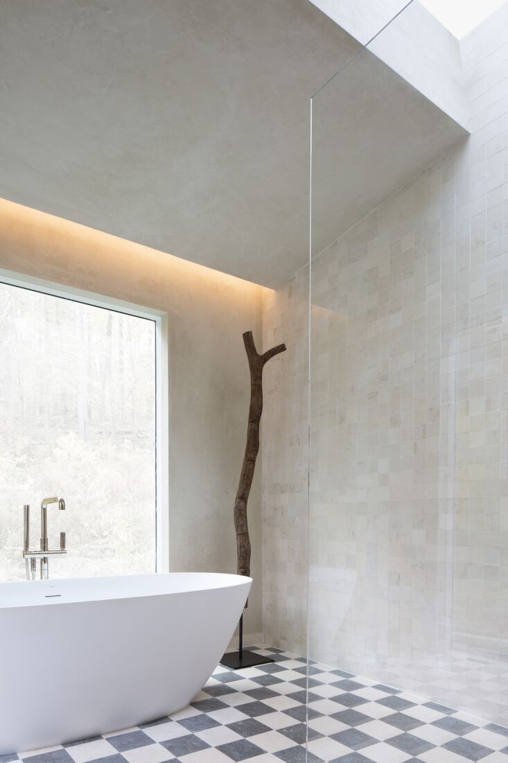 A freestanding white tub surrounded by patterned limestone tiles and large ribbed glass windows.