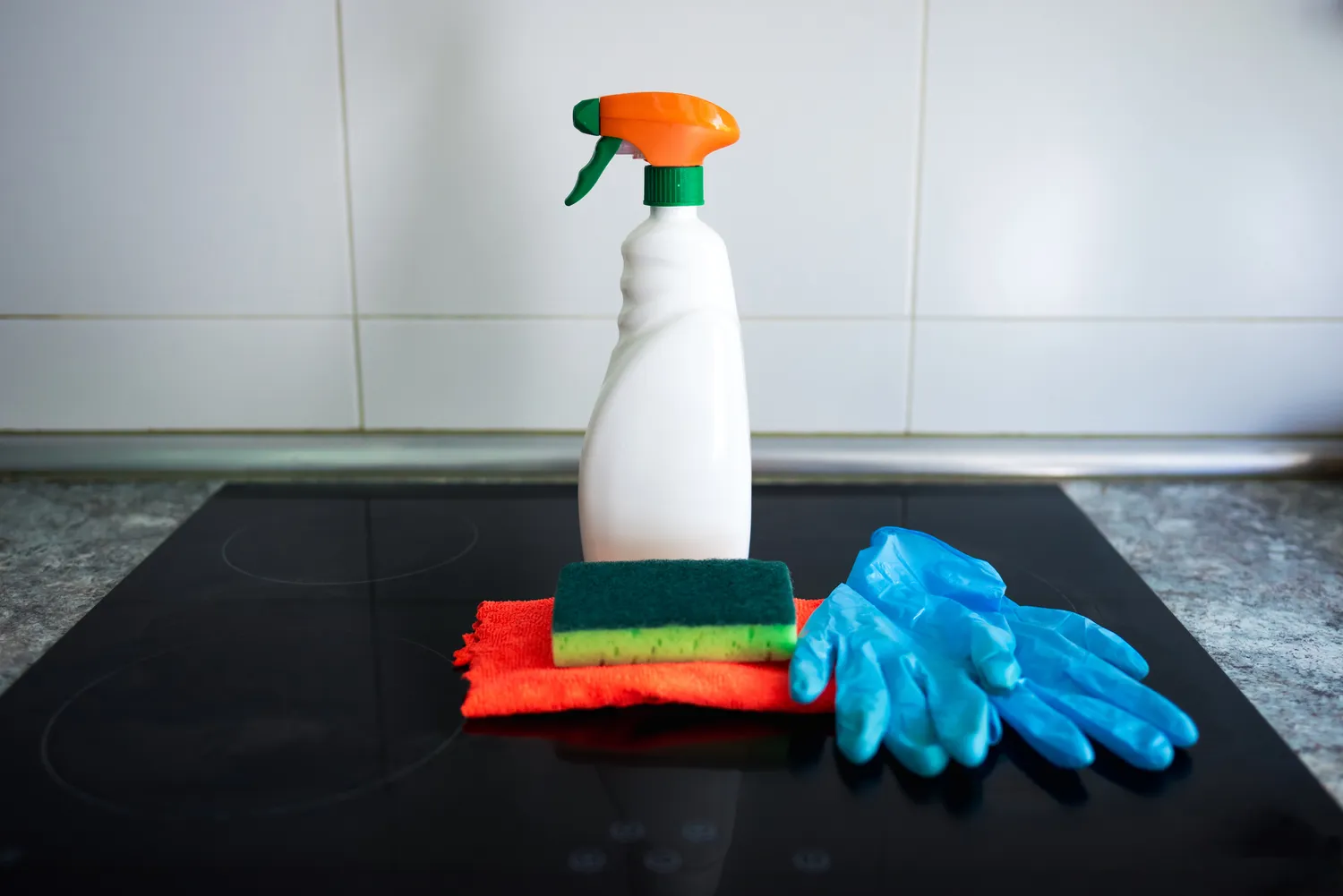Cleaning supplies like a spray bottle and sponge on a kitchen countertop next to an induction stove.