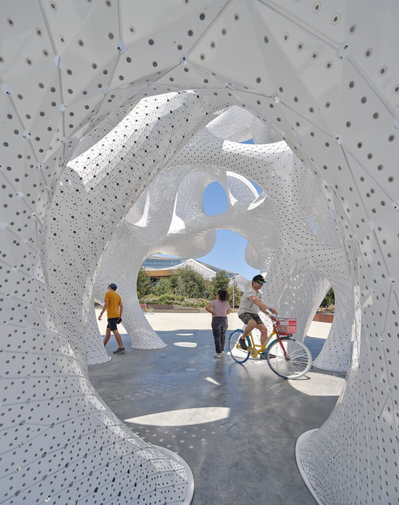 People walking and riding a bicycle through the large openings of the white sculptural pavilion on a sunny day.