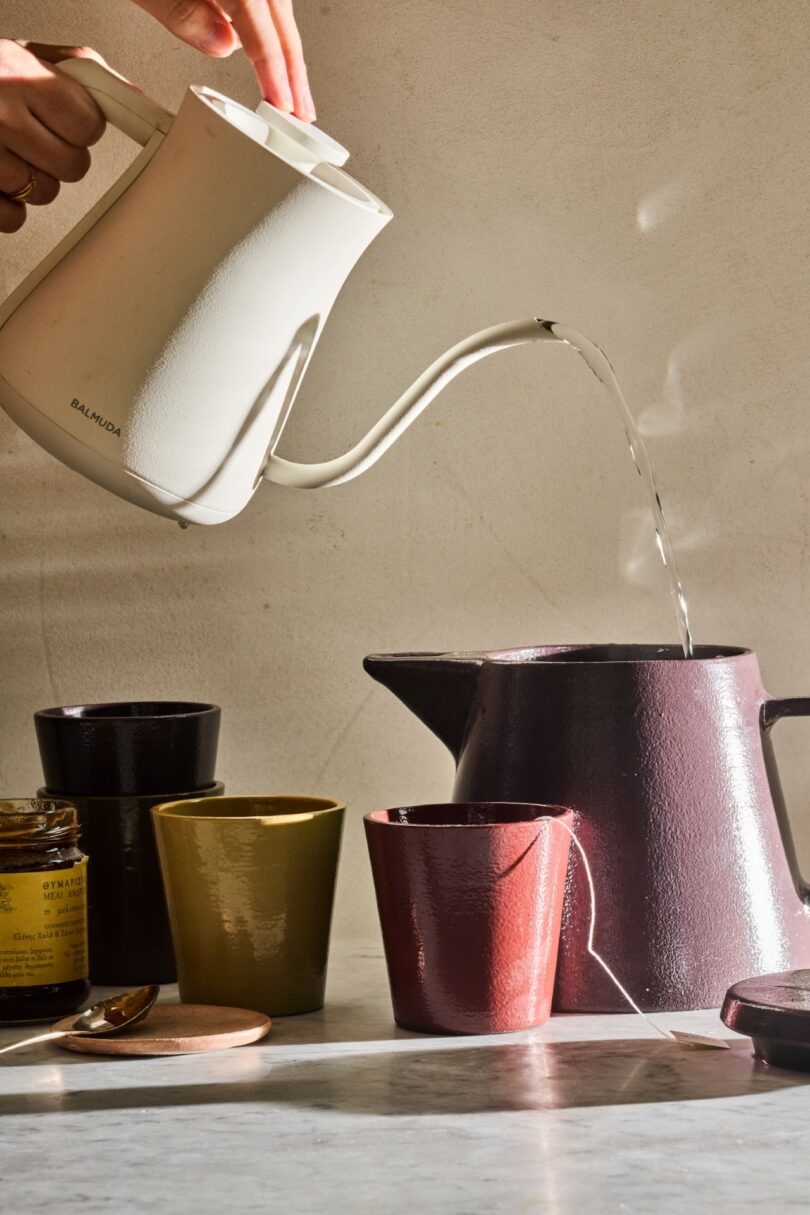 A hand pours hot water into a maroon teapot on a sunlit countertop with tea accessories.