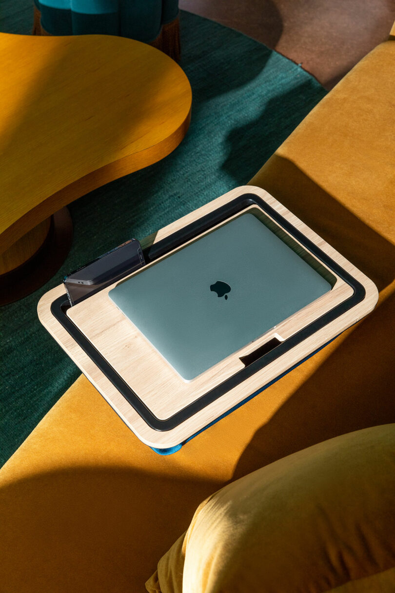 A closed laptop and phone resting on the wooden lap desk on a mustard yellow couch in a stylish living room.