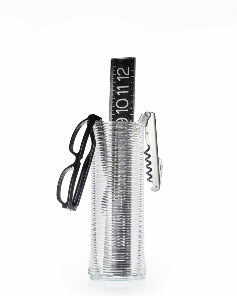 A clear ribbed glass vessel holding a ruler, eyeglasses, and a corkscrew.