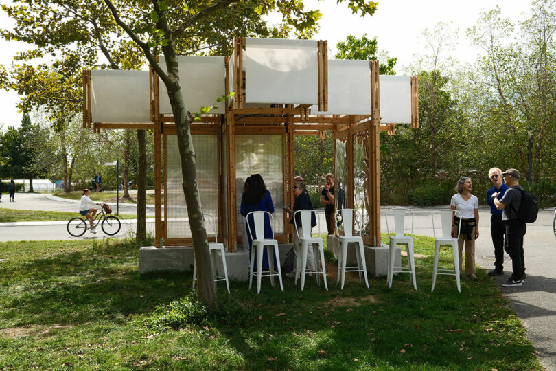 A wooden KlimaKover pavilion in a park with white cooling panels and people sitting nearby.