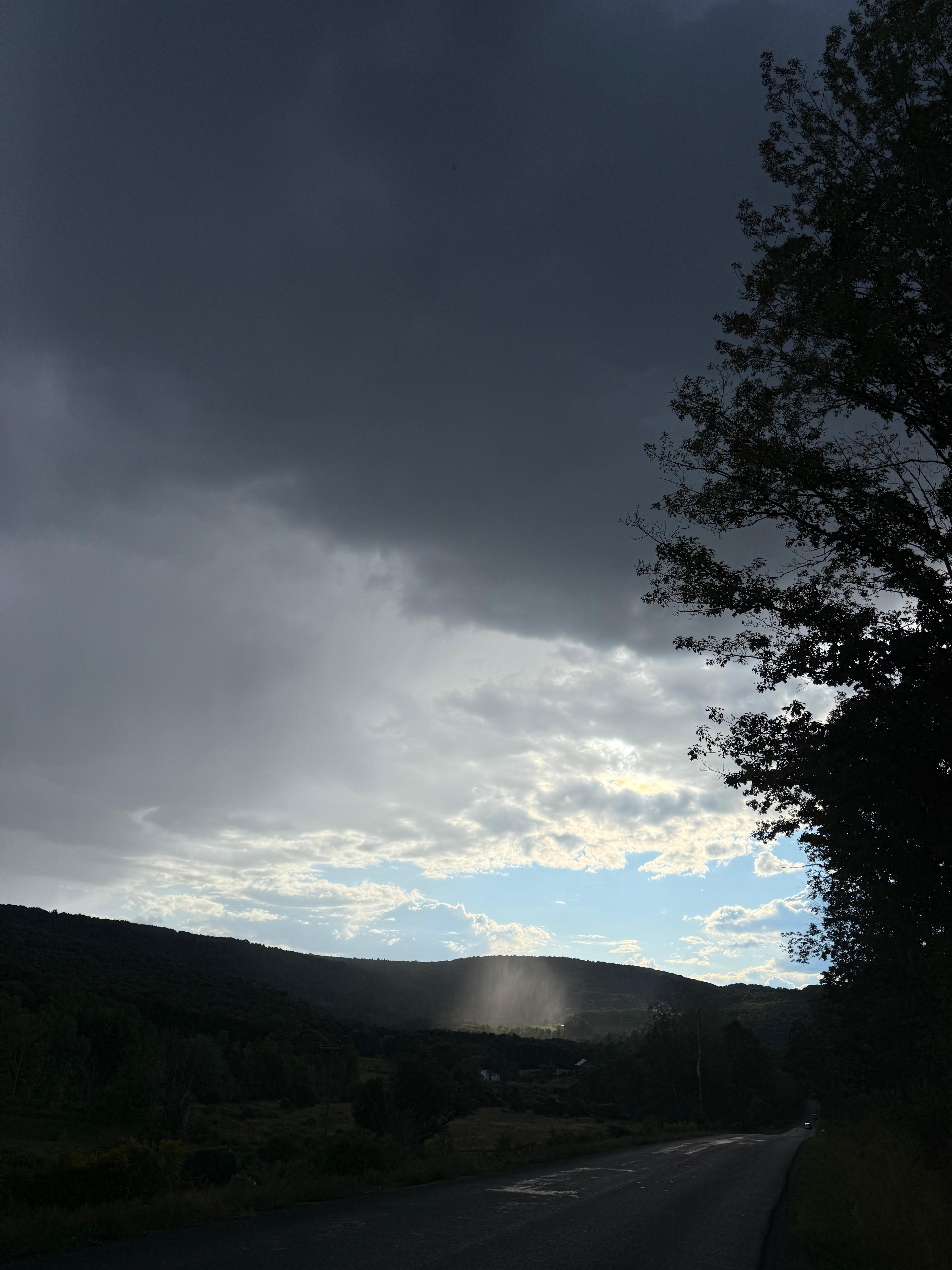 Dramatic dark clouds and a setting sun over a rural mountain landscape.