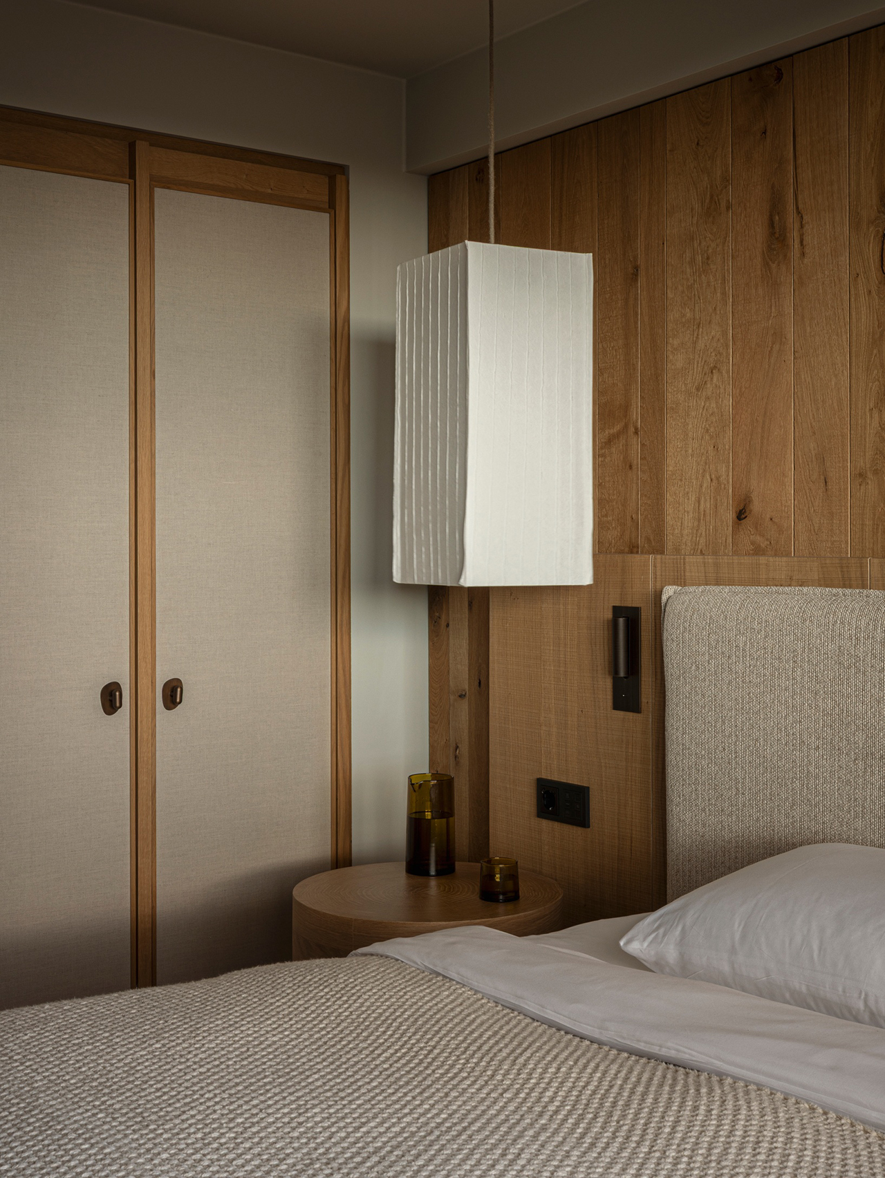 A modern guest bedroom featuring wooden paneling, a textured bed with cream linens, and a stylish pendant lamp.