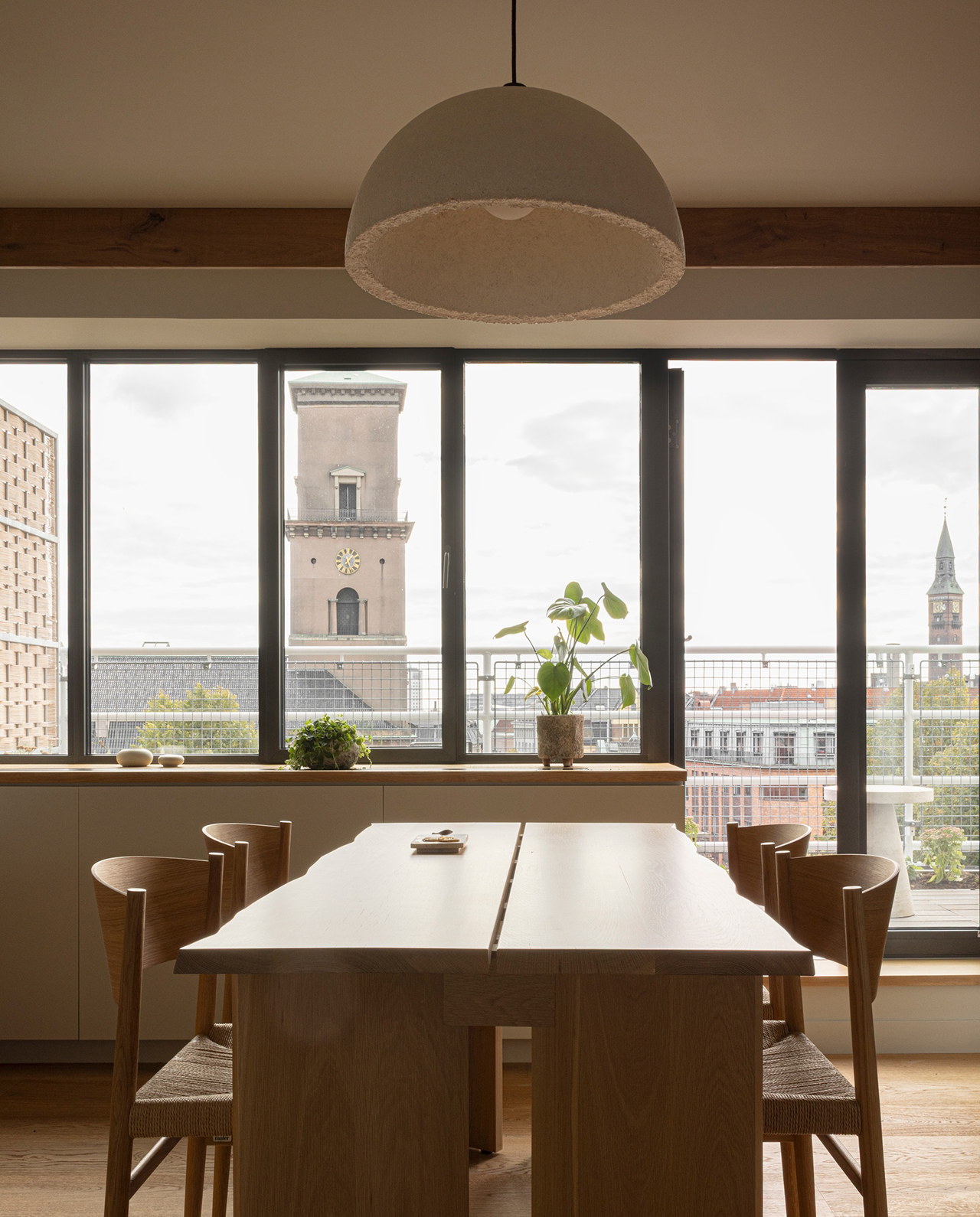A minimalist dining room with wooden furniture and potted plants, looking out through large windows onto historic Copenhagen architecture.