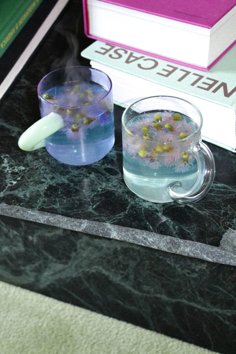 Two glass mugs with chamomile tea and flowers on a dark marble table next to books.