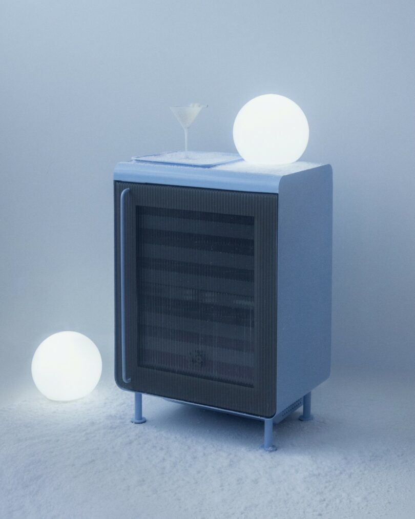 A modern blue refrigerator set on a snowy surface with a martini glass, evoking a sense of extreme cold.
