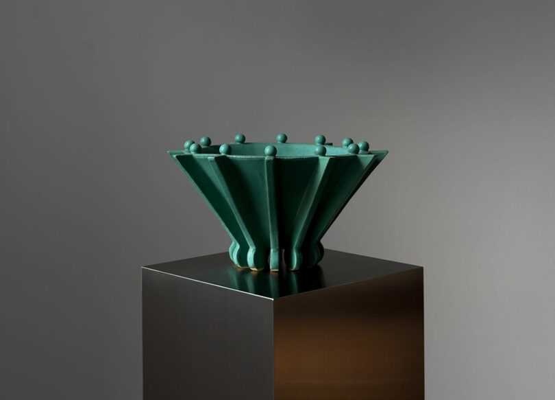 A green ceramic sculpture with a fluted, inverted cone shape on a reflective pedestal.
