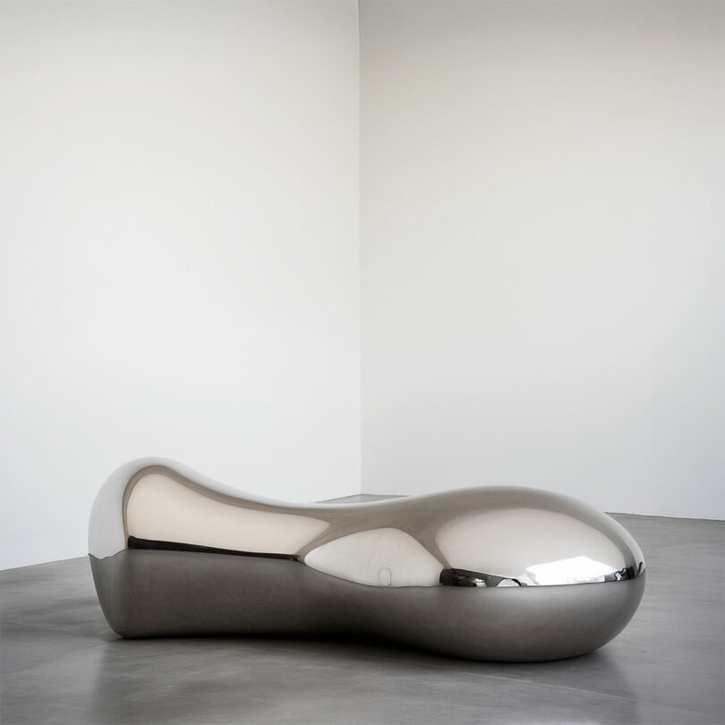 A large, smooth, bean-shaped metallic sculpture on a gray concrete floor in a minimalist gallery space.