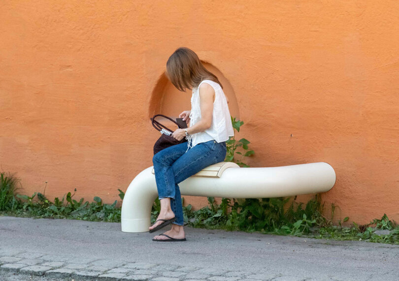A woman sitting on a vibrant orange curved pipe bench against a matching wall.