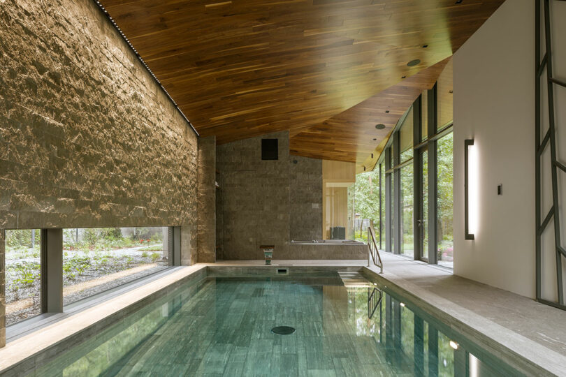 Indoor swimming pool with stone walls, wood-paneled ceiling, and floor-to-ceiling windows showing forest views.