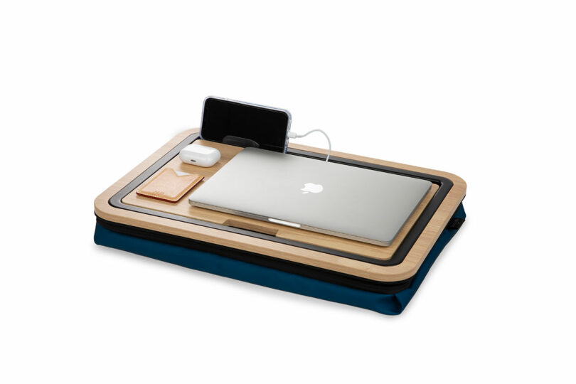 Close-up of the wooden lap desk surface holding a laptop, smartphone, and accessories with a blue cushion base.