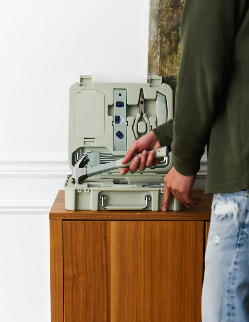 A person wearing a green jacket opens a mint green TINKR toolbox on top of a wooden cabinet.